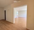 Empty room with light wooden flooring and white walls, featuring an open doorway leading to another room.