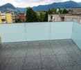Modern balcony with grey tiled flooring and glass railing, overlooking a cityscape with mountains in the background.