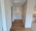 Bright hallway with wooden flooring, white walls, and built-in cabinets on one side. Features recessed lighting and a closed door at the end.