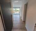 Modern hallway with wooden flooring, white walls, and recessed lighting. Features a large window at the end leading to a balcony or outdoor area.