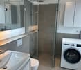 Modern bathroom with a white sink, toilet, glass-enclosed shower, and a private washing machine. Features include a large mirror and built-in cabinets.