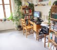 A well-lit study or home office with a wooden desk, computer setup, bookshelf filled with books, and several potted plants near the window.