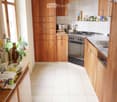 A modern kitchen with wooden cabinets, white tiled floor, and stainless steel appliances including a stove, oven, and sink. There are various kitchen items on the counter and a small plant near the window.