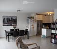 A modern, open-plan living and dining area with a dining table and chairs, a refrigerator, a bookshelf, and a comfortable armchair. The space features white walls, tiled flooring, and a large cityscape picture on the wall.