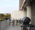 Balcony with a small table, chairs, and a barbecue, overlooking a cityscape with trees and buildings.