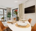 Dining area with a white table set for six, surrounded by orange chairs, a bottle of champagne, and decorative vases with flowers. Large windows offer a view of the city.