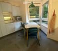 Bright kitchen with white cabinets, stainless steel oven, microwave, and a dining table with chairs, including a baby chair. Large windows provide natural light.