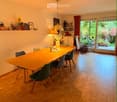 Dining room with a long wooden table surrounded by green chairs, a hanging pendant light, and a shelf with decorative items.