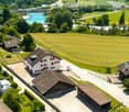 Aerial view of a rural residential area with multiple houses, a parking lot, and surrounding green fields and a river.