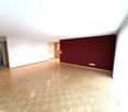 Empty room with wooden herringbone flooring, white walls, and a dark red accent wall. Features include a doorway to another room and a partially visible bathroom with tiled walls.