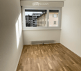 Empty room with wooden flooring, white walls, and a large window showing a cityscape view. No furniture or equipment present.