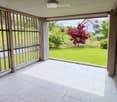 Spacious balcony with white tiled flooring, wooden railings, and large glass panels offering views of a well-maintained garden and surrounding houses.