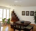 Bright living and dining area with large windows and sliding doors leading to a balcony. Features a grey sofa, coffee table, dining table with chairs, a wooden cabinet, decorative vases, picture frames, and a potted plant.