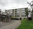 Modern multi-story building with large windows and a playground area in front, featuring wooden play structures and a slide.