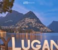 Scenic view of Lake Lugano with illuminated town and mountains in the background, featuring large white letters spelling "MY LUGANO" in the foreground.