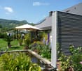 Well-maintained garden with a pond, various plants, flowers, and a modern house in the background. Features include a patio umbrella, decorative statues, and a scenic mountain view.