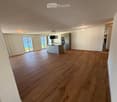 Open-plan living and kitchen area with wooden flooring, modern kitchen island, refrigerator, oven, and television. Large windows offer a view of the water.