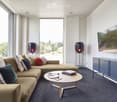 Modern living room with large windows, grey carpet, beige sectional sofa, wooden coffee table, floor lamps, and a flat-screen TV mounted on the wall.