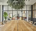 A modern, well-lit room with large glass partitions, a long wooden table surrounded by chairs, and a hanging plant. The space features contemporary decor, including a mirror and a screen in the background.