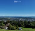 Panoramic view of a town and surrounding landscape from a hilltop, with houses, trees, and distant mountains visible.