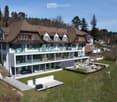 A large, two-story house with a modern extension featuring large glass windows and a spacious balcony. The traditional section has a pitched roof and green shutters. The property includes a well-maintained lawn, patio area, and surrounding trees.