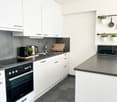 Modern, minimalist kitchen with white cabinets, gray countertops, built-in appliances including a dishwasher and microwave, and decorative plants and kitchen accessories on the shelves.