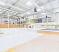 Large open warehouse space with high ceilings, white walls, and yellow accents. The space has a concrete floor with yellow markings, and there are various storage racks and equipment visible.