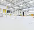 Large open commercial space with high ceilings, white walls, and yellow accents. The space has a stage area and appears to be set up for events or performances. There are also some storage areas and parking spaces visible.