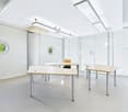 Bright and modern office space with large windows, white walls, and minimalist furniture including desks and chairs. The room has recessed lighting and a clean, professional atmosphere.