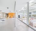 Large open-plan office space with concrete floors, white walls, and glass partitions. The space features a seating area with yellow chairs and a table, as well as a raised platform with additional seating. The ceiling is high and has exposed beams and lig