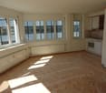 Empty room, hardwood floor, large windows, kitchen area with oven and sink, radiators, wooden cabinets