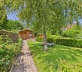 Path leading to wooden house, surrounded by hedges, lounge chairs, green grass, trees, house in the background