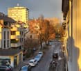 A street lined with cars parked on either side, buildings, a tree, and a balcony with plants