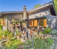 detached house, wooden exterior, yellow shutters, chimney, outdoor dining area with table and chairs, garden with various plants and shrubs, brick pathway, clear blue sky