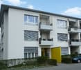 4-story building, white exterior, windows, yellow front door, car parked in front, hedges