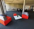 modern office with gray carpet, red and gray chairs, white table, small potted plant, large windows, tables and chairs in the background