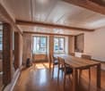 This image shows a combined living and dining room area with a rustic, cozy feel. The room features exposed wooden beams on the ceiling, wooden floors, and large windows that let in natural light. The dining table and chairs are made of wood, and there is