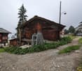 wooden building on slope, two smaller houses, greenery around, dirt path in front