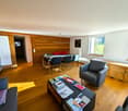 Spacious living room with wooden walls, hardwood floors, large windows, dining table, sofa, armchair, and a fireplace