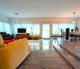 Spacious open-plan living area with large windows, hardwood floors, yellow and black sofas, TV stand, dining table and chairs, and a raised platform with glass doors leading to a balcony or terrace.