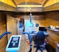 The image shows a small, cozy home office space inside a wooden cabin or shed. The room has a slanted wooden ceiling and walls, with a desk setup that includes a computer monitor, keyboard, and a blue exercise bike. There are various personal items and of