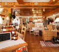 A restaurant interior with wooden ceiling, wooden floor, wooden tables, wooden chairs, white tablecloths, jars of honey, blackboard with text, wooden wine cabinet, white lamps, shelves with bottles, bottles, a gas stove, a plant on the wall, and a cabinet