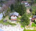 Aerial view of a two-story wooden chalet-style house surrounded by lush greenery, trees, and a winding road. The house has a balcony or terrace and appears to be situated on a raised ground floor. There are additional buildings and houses visible in the b