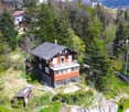 2-story chalet-style house with a red roof, surrounded by lush greenery and trees, situated on a hillside with a view of the mountains and valley below