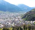 Aerial view of a mountainous city with a mix of high-rise buildings, residential areas, and green spaces. The city is surrounded by towering mountains and lush vegetation. There appears to be a river or stream running through the center of the city.