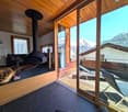 Cozy living room with wooden ceiling, large windows overlooking snowy mountains, a black wood-burning stove, and a balcony/terrace with outdoor furniture.