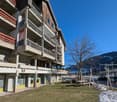 Multi-story building with wooden and concrete exterior, balconies on each floor, ground floor with retail spaces, surrounded by snow-covered grass and trees
