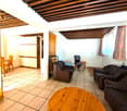 Spacious living room with wooden beamed ceiling, tile flooring, leather sofa and armchair, wooden coffee table, and a desk with a chair. There appears to be a balcony or terrace visible through the window.
