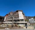 Multi-story apartment building with balconies, surrounded by trees and mountains in the background.