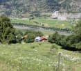 The image shows a picturesque rural landscape in a mountainous region. In the foreground, there is a grassy field with wildflowers, and in the background, there are several houses or cabins nestled among the trees. The landscape is dominated by rolling hi